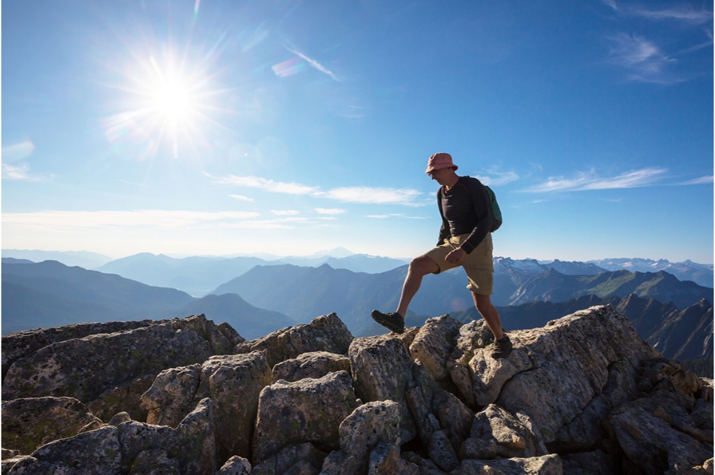 un randonneur grimpe sur des rochers dans les montagnes. Il est équipé pour l'été avec un short de randonnée, un tee-shirt lanche longue, des chaussures de randonnée, un sac à dos et un bob pour le soleil.