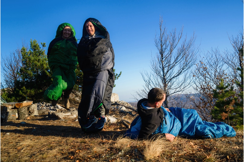 dans une forêt, 3 randonneurs sont dans leur sac de couchage. L'un est couché par-terre, les 2 autres sautent avec leur sac de couchage face au photographe en souriant.