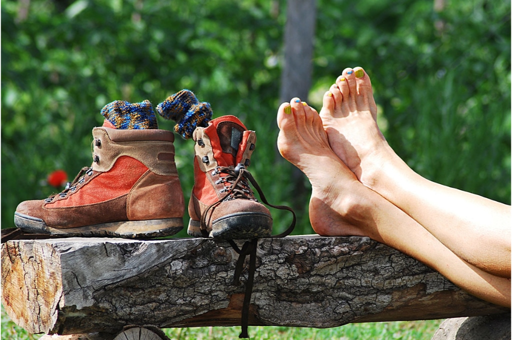 2 jambes croisées vernies sont posée sur un banc fait avec un tronc d'arbre coupé en 2. à côté, 2 chaussures de randonnée marron et oranges sont posées avec des chaussures colorées à l'intérieur.