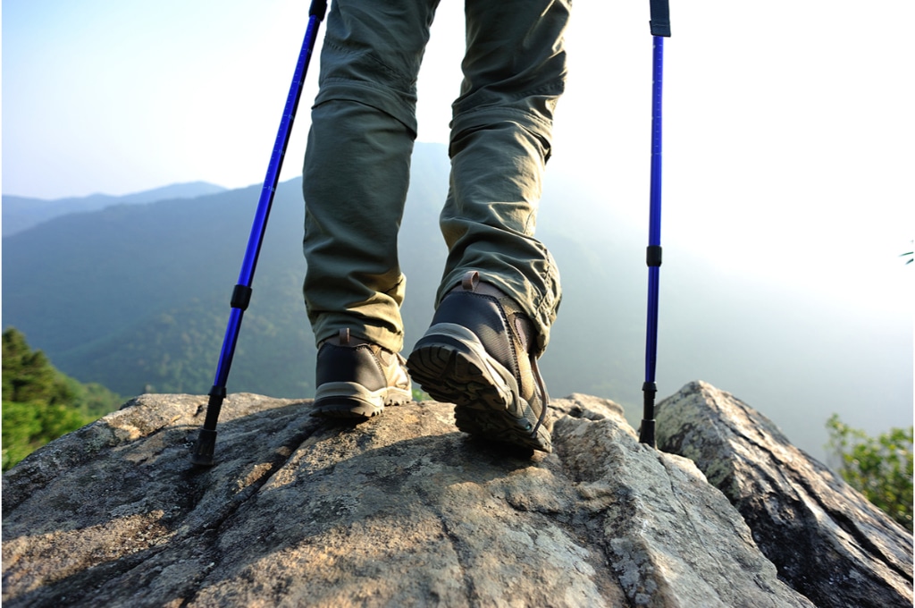vue sur les jambes d'un randonneur de montagne avec des bâtons de randonnée. il est au bord d'une falaise, entouré de montagnes. il porte des chaussures de randonnée et un pantalon de randonnée.