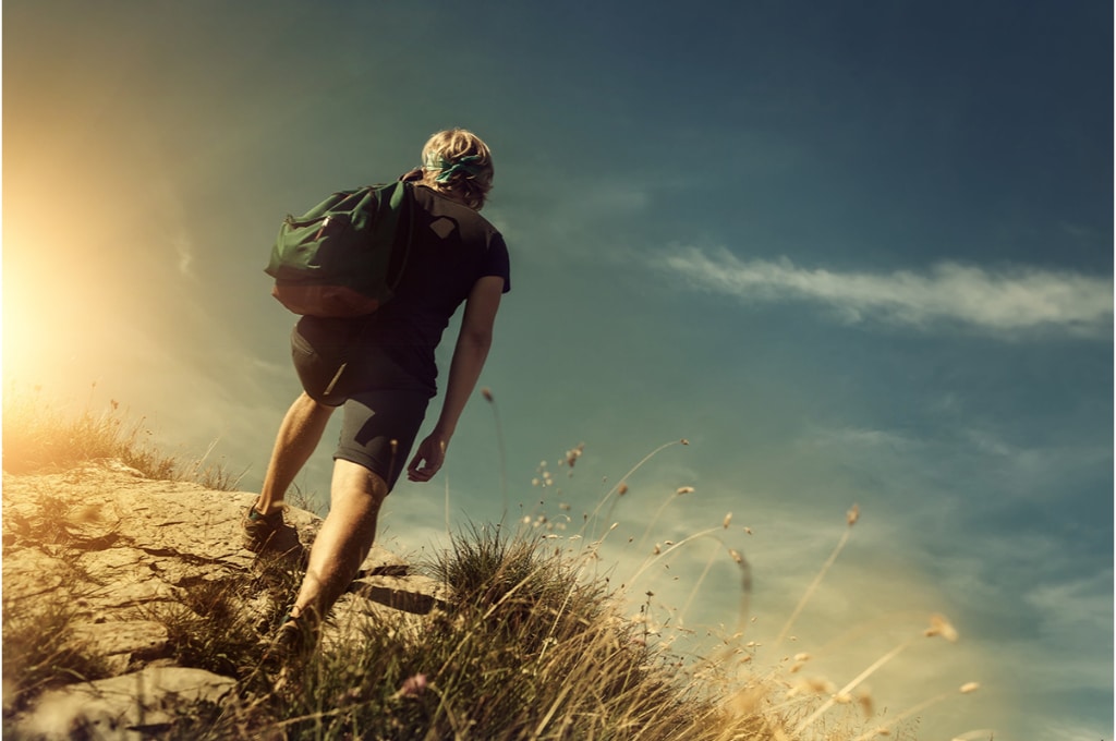 un homme blond monte une colline avec son sac à dos vert dans le dos. il fait beau ciel bleu. ses cheveux sont attachés. il est en short et tee-shirt manches courtes.