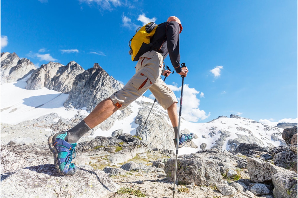 un randonneur marche dans des montagnes enneigées sous un beau ciel bleu. il porte des bâtons de randonnée et de bonnes chaussures de randonnée. il porte un short et un haut manche longue ainsi qu'un sac à dos. sa tête est protégée grâce à un bob rose.