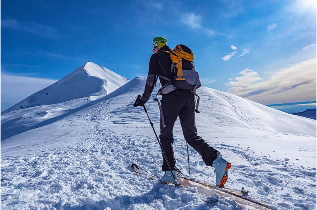 sur le sommet d'une montagne enneigée, une personne fait du ski de fond de randonnée avec des bâtons. elle porte un sac à dos et une foulard autour de la tête.
