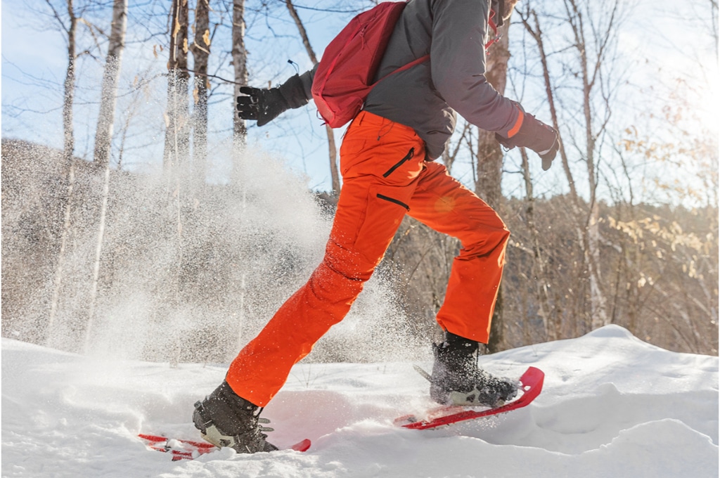 dans la neige dans une forêt, un homme fait des raquettes. il porte un pantalon de randonnée orange. il a des gants et un sac à dos rouge.