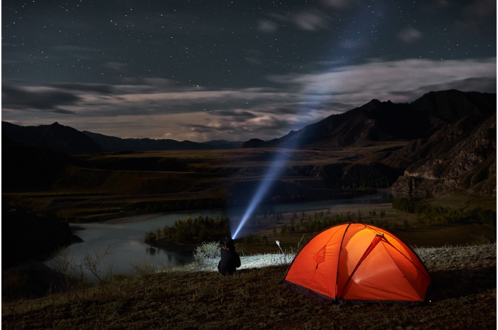 dans la nature en pleine nuit, un randonneur dans une tente orange éclairée, éclaire le ciel avec une lampe torche