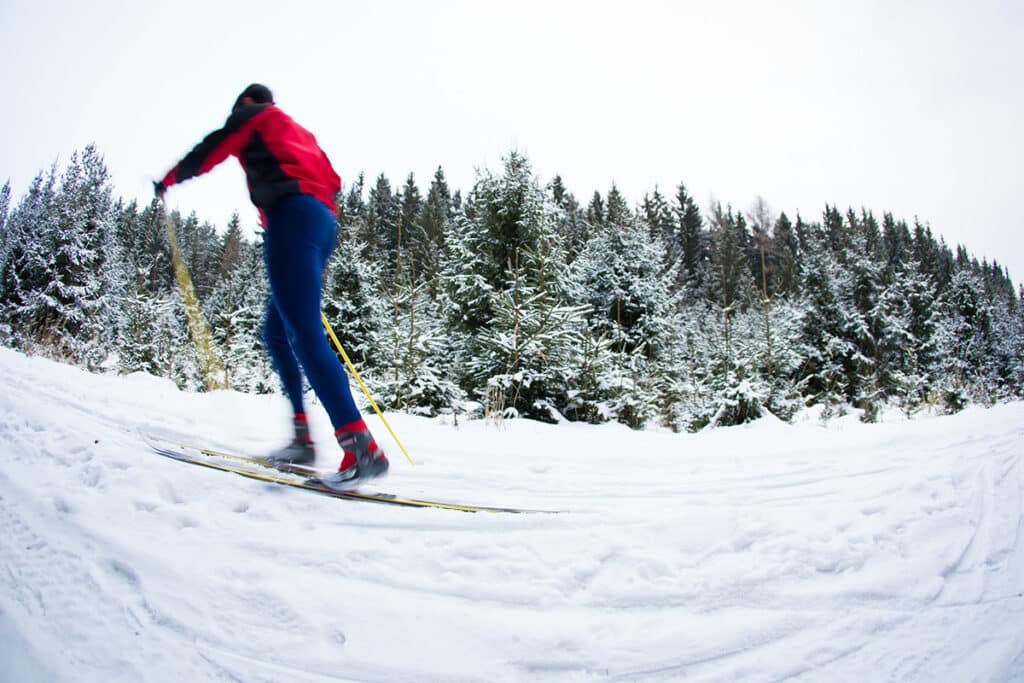vue sur une forêt derrière une piste de ski. un randonneur à ski passe sur le neige. il porte des skis et des bâtons de ski jaunes.