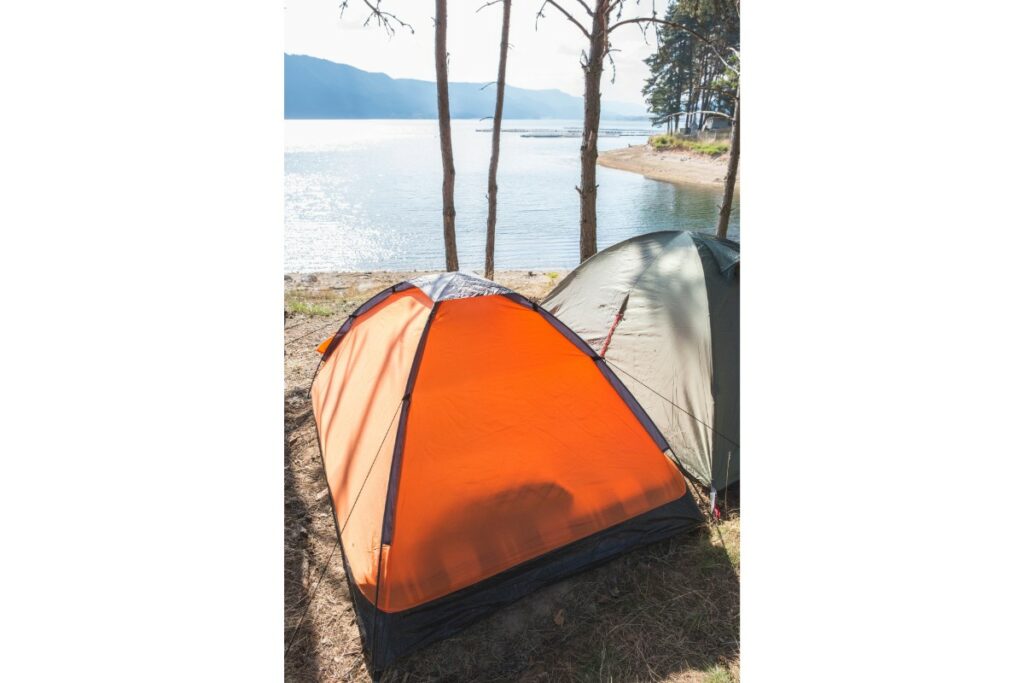 2 tentes, une orange et une grise sont plantées, côte à côte devant une étendue d'eau dans une forêt.