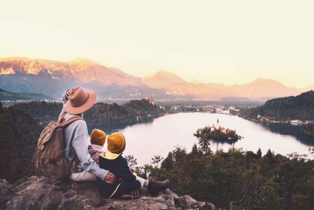 devant un paysage de montagne et de lac, une personne est assise avec ses 2 enfants en haut d'un rocher. ils contemplent le paysage.