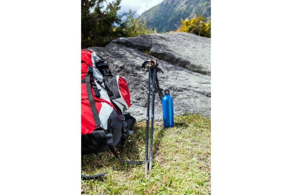 en montagne, 2 batons de randonnée sont posés sur un rocher. une gourde bleue est posée à côté ainsi qu'un sac à dos de randonnée rouge noir et gris.