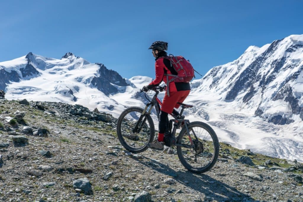 devant un paysage de montagnes enneigées, un cycliste randonne sur son vélo. Il est équipé de la tête au pied en rouge avec un sac à dos rouge et un casque à vélo.