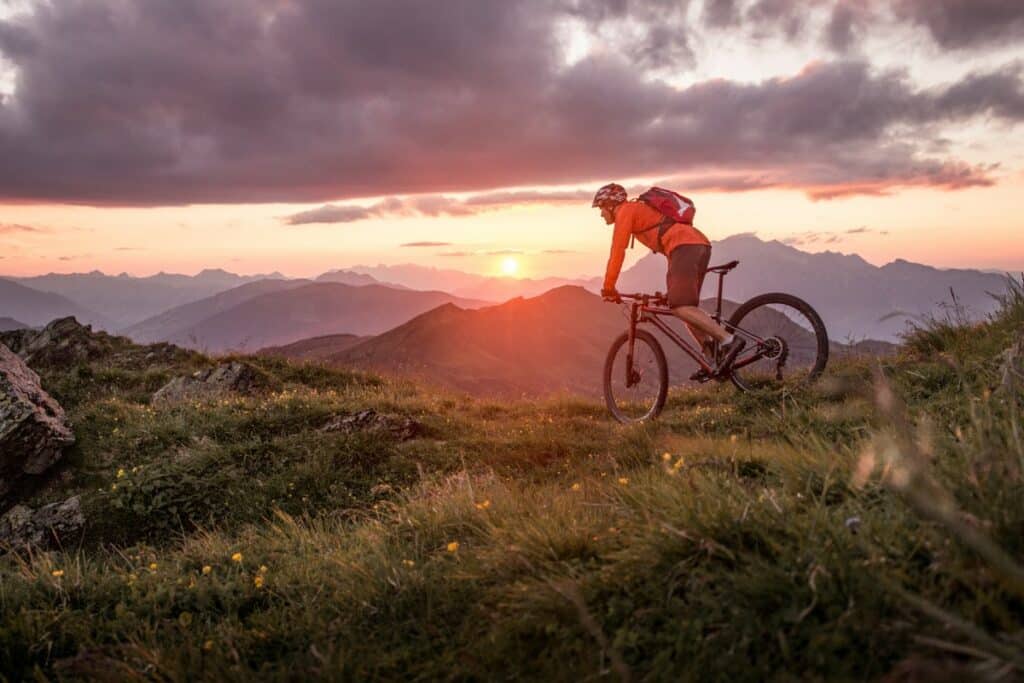 devant un paysage de montagnes, au lever du soleil, un randonneur à vélo pédale sur son vélo. il porte un casque de vélo, un sac à dos rouge, un short foncé et un haut à manches longues orange.