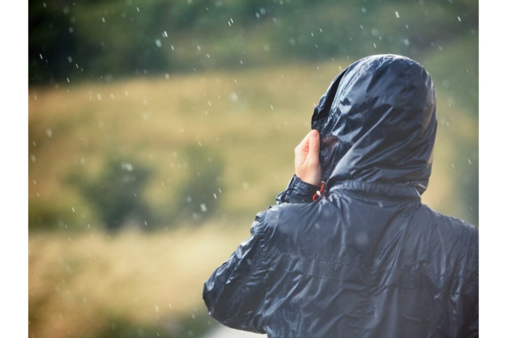 sous la pluie, une personne de dos porte une veste de pluie avec une capuche sur la tête. elle tient la capuche avec sa main gauche.
